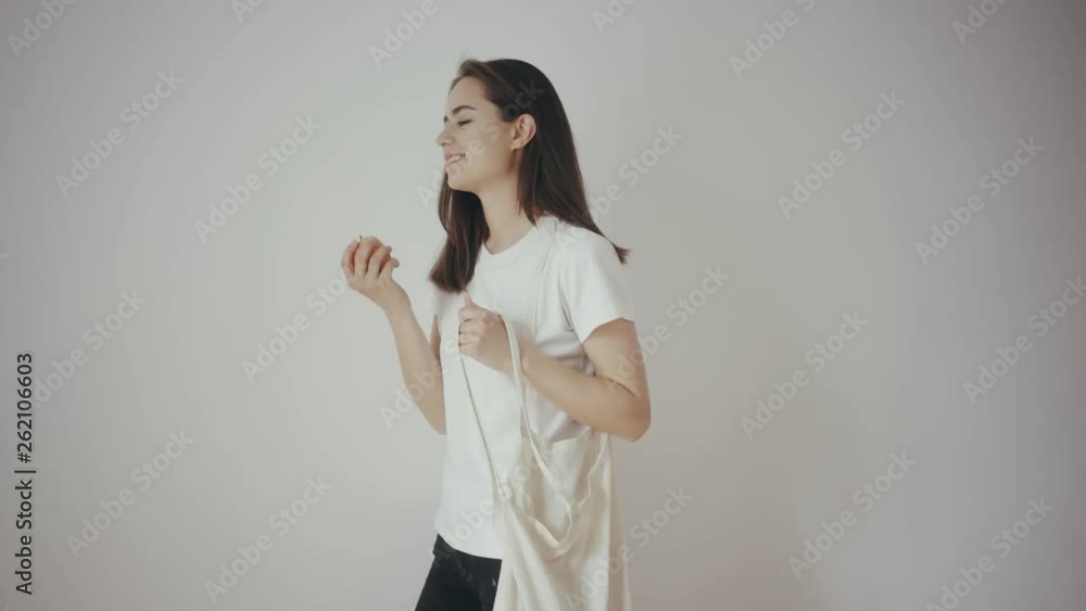 Young hipster smiling girl with white blank cotton shopper tote bag puttin an apple inside, mock-up of blank bag, white wall on the background, concept of shopping Young hipster smiling girl with white blank cotton shopper tote bag puttin an apple inside, mock-up of blank bag, white wall on the background, concept of shopping