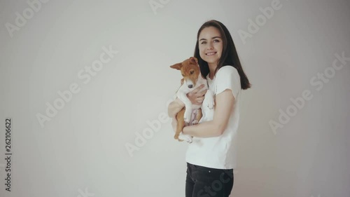 Cute young hipster girl hugging and kissing lovely basenji puppy dog. Girl wearing blank white cotton t-shirt standing in front of white isolated wall. Concept of love and care between pet and owner