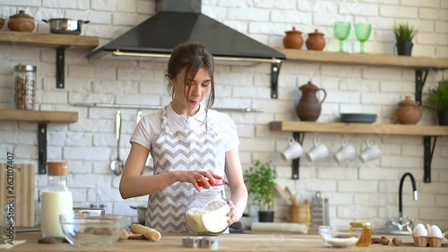 Mother is cooking at the kitchen. Preparin dough, holding bottle of flour. While her little daughter playing hide and seek and taking oatmeal cookies from the table. photo