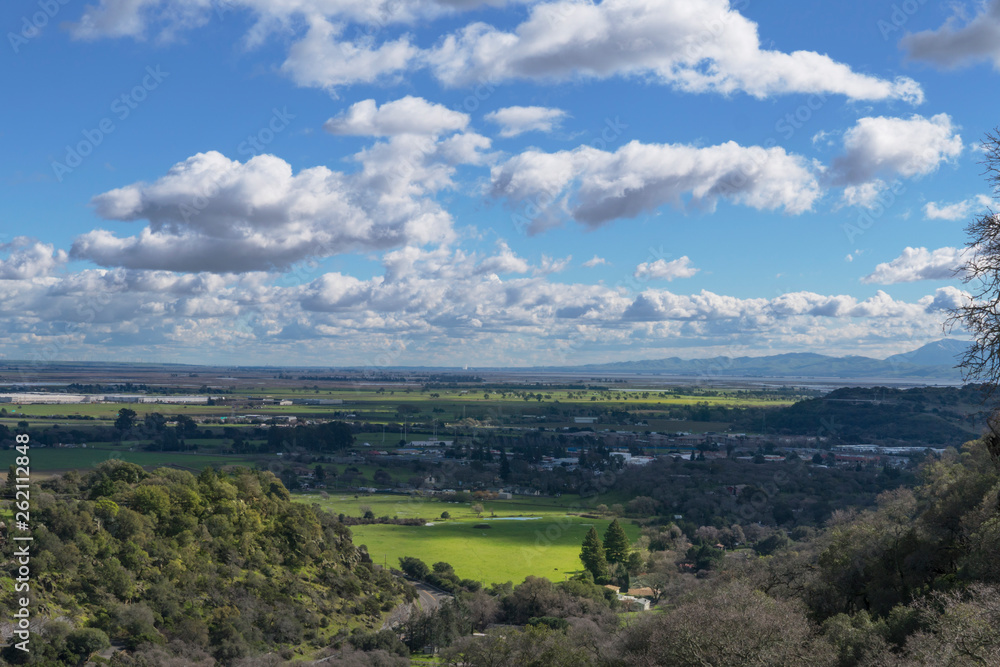Landscape view from mountaintop, green sunlit valley with blue sky and white clouds, farmland Northern California wine country.