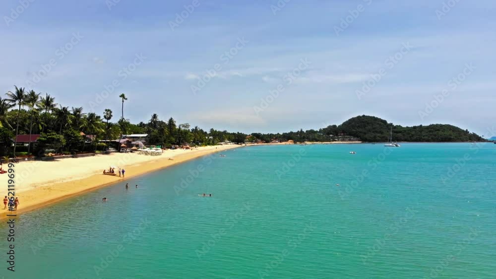 Wide, beautiful sandy beach and calm ocean with shades of blue as people enjoy the warm sunshine and water of a tropical paradise. Scenic island beauty of Thailand. Drone flying low over the sea. Wide, beautiful sandy beach and calm ocean with shades of blue as people enjoy the warm sunshine and water of a tropical paradise. Scenic island beauty of Thailand. Drone flying low over the sea.