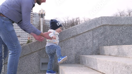 bearded man in sunglasses holding his little son by the hand helps the baby boy to climb the stairs in the city park on sunny spring day background in slow motion middle shot 4K video