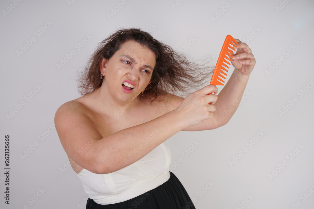 emotional unhappy woman with disheveled hair tries to comb them and suffers. curvy girl stands on white background in Studio alone. emotional unhappy woman with disheveled hair tries to comb them and suffers. curvy girl stands on white background in Studio alone.