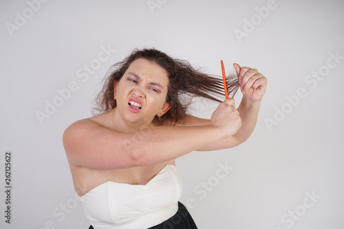 emotional unhappy woman with disheveled hair tries to comb them and suffers. curvy girl stands on white background in Studio alone.