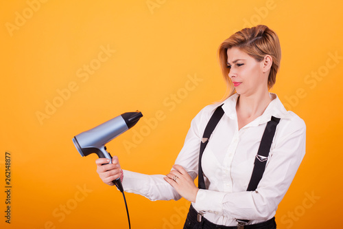 Pretty young woman looking confused at her hair drayer over yellow background.