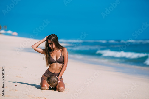 Sexy tanned model with sunglasses in brown swimsuit with coconut posing on white sandy beach
