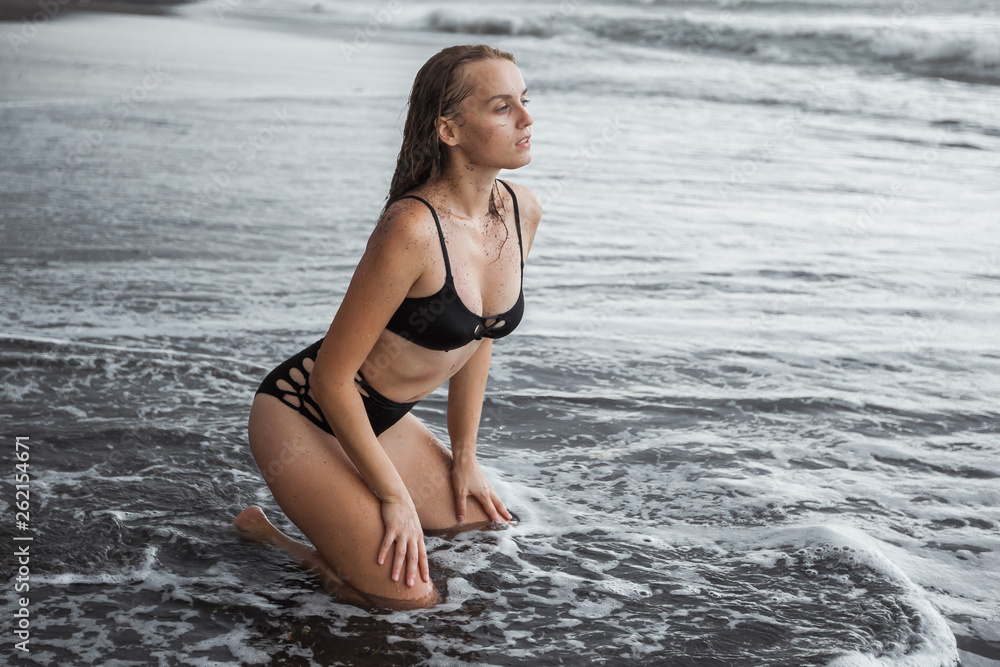 The girl in the black swimsuit on the black sand is kneeling.