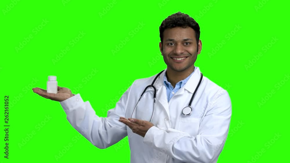 Young male doctor presenting pills on green screen. Smiling Indian doctor with stethoscope showing bottle of tablets. Chroma Key background. Young male doctor presenting pills on green screen. Smiling Indian doctor with stethoscope showing bottle of tablets. Chroma Key background.