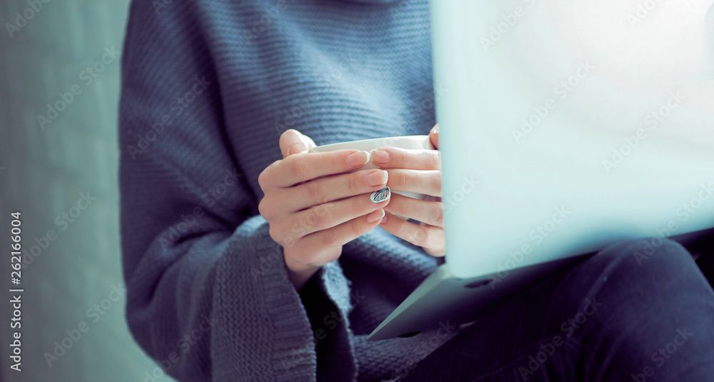 Girl sending a message, e-mail, woman typing something on the laptop in her lap sitting near window and holding cup of coffee close-up.
