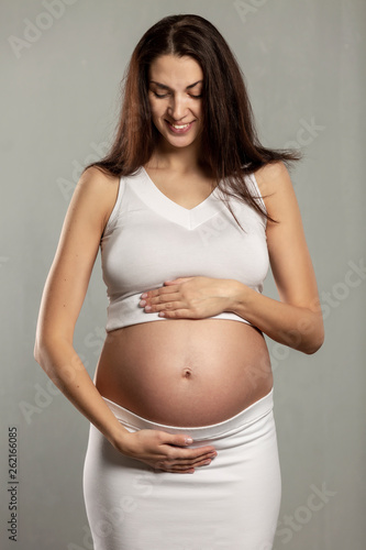 Young beautiful pregnant woman gently hugs belly and smiles, vertical, gray background Free Download Young beautiful pregnant woman gently hugs belly and smiles, vertical, gray background - GFXLoad