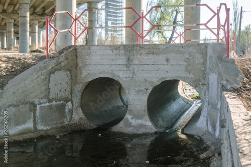 Two concrete sewer pipe covered under bridge. Black dirty water.