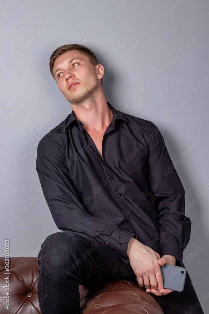 Attractive young handsome man wearing black shirt sitting on the leather sofa holding smartphone. Comfort and relaxation. Attractive young handsome man wearing black shirt sitting on the leather sofa holding smartphone. Comfort and relaxation.