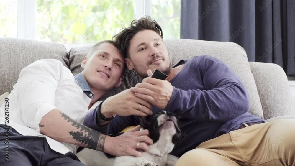Young gay male couple relaxing on couch with dog in living room. Caucasian, european men sitting together on sofa. Kissing. Happy gay friendship, relationship, lifestyle concept. Young gay male couple relaxing on couch with dog in living room. Caucasian, european men sitting together on sofa. Kissing. Happy gay friendship, relationship, lifestyle concept.