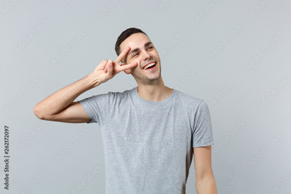 Portrait of cheerful smiling young man in casual clothes looking aside, showing victory sign isolated on grey wall background in studio. People sincere emotions, lifestyle concept. Mock up copy space. Portrait of cheerful smiling young man in casual clothes looking aside, showing victory sign isolated on grey wall background in studio. People sincere emotions, lifestyle concept. Mock up copy space.