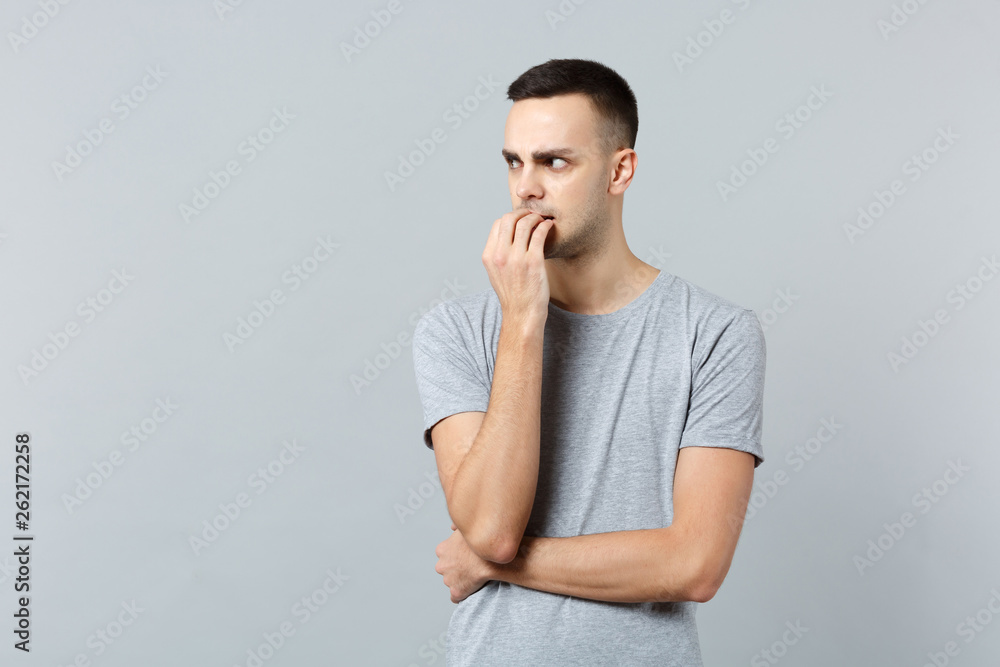 Portrait of nervous young man in casual clothes gnawing nails looking aside isolated on grey wall background in studio. People sincere emotions lifestyle concept. Mock up copy space. Advertising area. Portrait of nervous young man in casual clothes gnawing nails looking aside isolated on grey wall background in studio. People sincere emotions lifestyle concept. Mock up copy space. Advertising area.