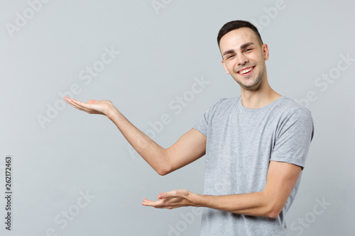 Portrait of cheerful funny young man in casual clothes looking camera, pointing hand aside isolated on grey wall background in studio. People sincere emotions, lifestyle concept. Mock up copy space.