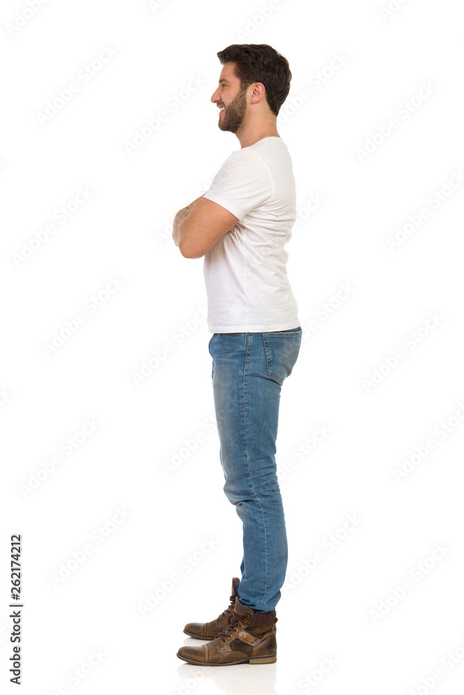 Smiling Man In Jeans And White T-shirt Is Standing With Arms Crossed. Side View. Smiling Man In Jeans And White T-shirt Is Standing With Arms Crossed. Side View.