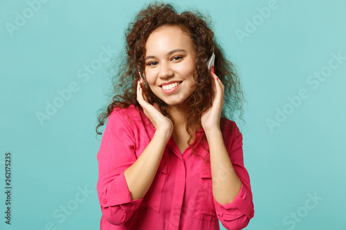 Smiling african girl in casual clothes looking camera, listening music with headphones isolated on blue turquoise background in studio. People sincere emotions, lifestyle concept. Mock up copy space. © ViDi Studio