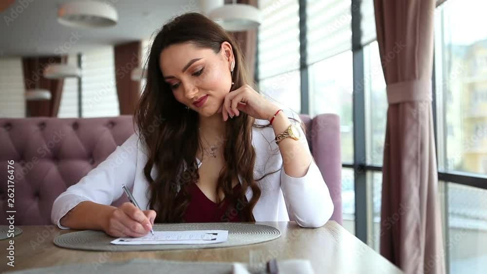 Cute nice girl sitting at a table in a cafe.The girl writes information on a sheet of paper. Cute nice girl sitting at a table in a cafe.The girl writes information on a sheet of paper.
