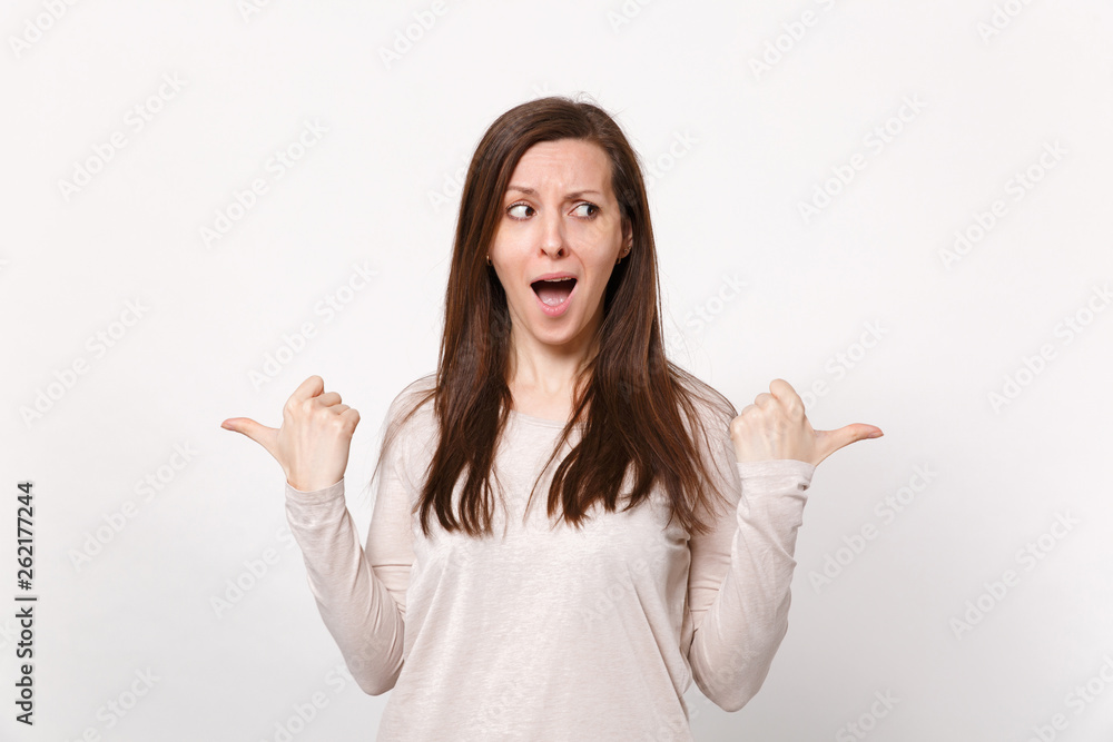 Portrait of excited amazed beautiful young woman in light clothes pointing thumbs aside isolated on white wall background in studio. People sincere emotions, lifestyle concept. Mock up copy space. Portrait of excited amazed beautiful young woman in light clothes pointing thumbs aside isolated on white wall background in studio. People sincere emotions, lifestyle concept. Mock up copy space.