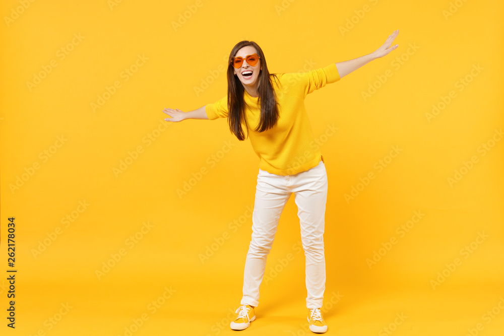 Laughing young woman in casual clothes, heart glasses standing and spreading hands isolated on yellow orange wall background in studio. People sincere emotions, lifestyle concept. Mock up copy space.