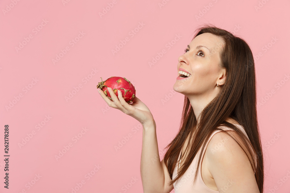 Side view of young woman looking up hold in hand fresh ripe pitahaya, dragon fruit isolated on pink pastel wall background in studio. People vivid lifestyle relax vacation concept. Mock up copy space. Side view of young woman looking up hold in hand fresh ripe pitahaya, dragon fruit isolated on pink pastel wall background in studio. People vivid lifestyle relax vacation concept. Mock up copy space.