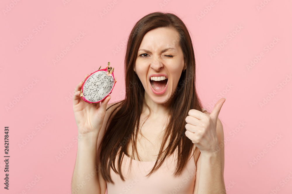 Crazy blinking young woman showing thumb up, holding half of fresh ripe pitahaya, dragon fruit isolated on pink pastel background. People vivid lifestyle relax vacation concept. Mock up copy space.