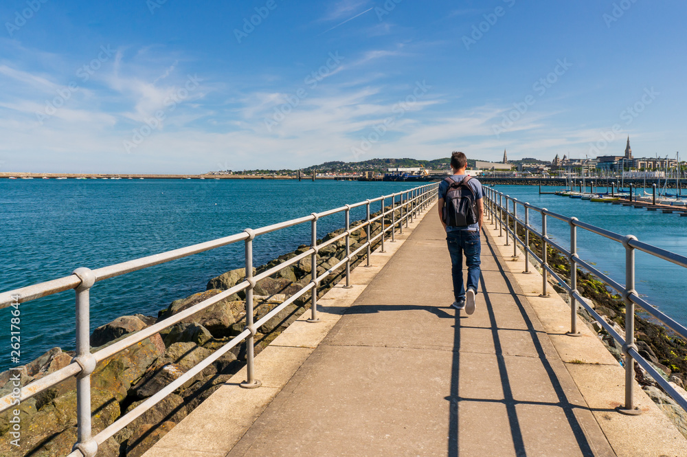 Young man walking on the Dun Laoghaire West Pier breakwater, in Dublin, Ireland and enjoying the lovely view of the village.  Hot weather on a beautiful Irish summer day.