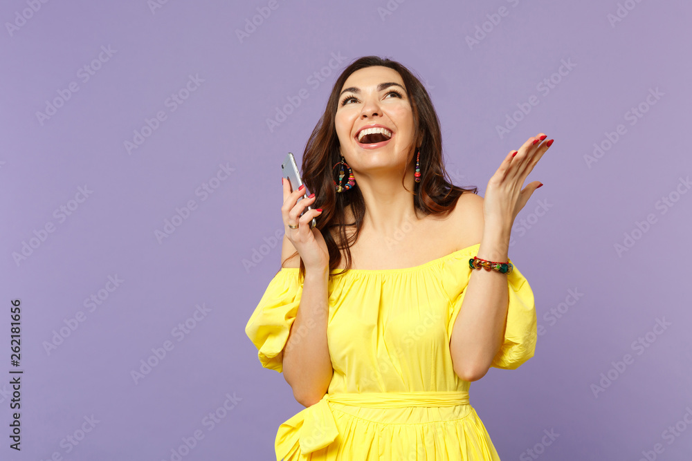 Fototapeta premium Cheerful young woman in summer dress using mobile phone, typing sms message, spreading hands, looking up isolated on violet background. People sincere emotions, lifestyle concept. Mock up copy space.