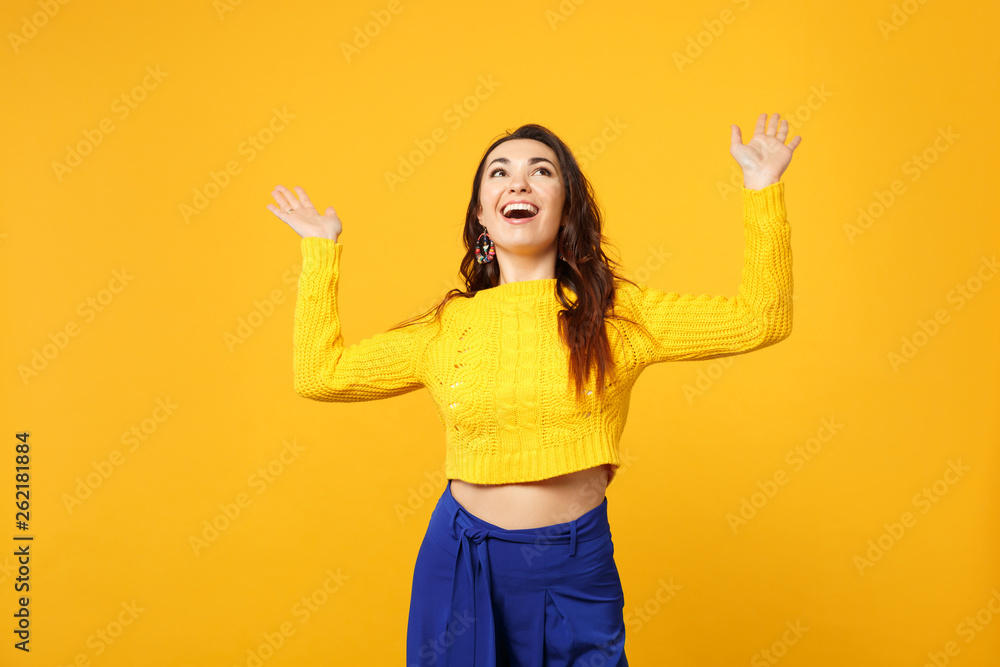 Portrait of cheerful young woman in sweater, blue trousers looking up, rising spreading hands isolated on yellow orange wall background. People sincere emotions lifestyle concept. Mock up copy space. Portrait of cheerful young woman in sweater, blue trousers looking up, rising spreading hands isolated on yellow orange wall background. People sincere emotions lifestyle concept. Mock up copy space.