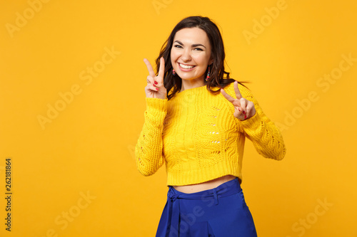 Portrait of cheerful young woman in sweater, blue trousers looking camera showing victory sign isolated on yellow orange wall background. People sincere emotions lifestyle concept. Mock up copy space.