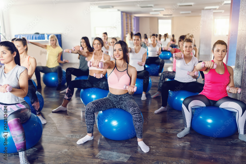 Group of focused and inspired group of sporty women sitting on pilates ball and lifting dumbbells in gym. Sweat, smile and repeat.
