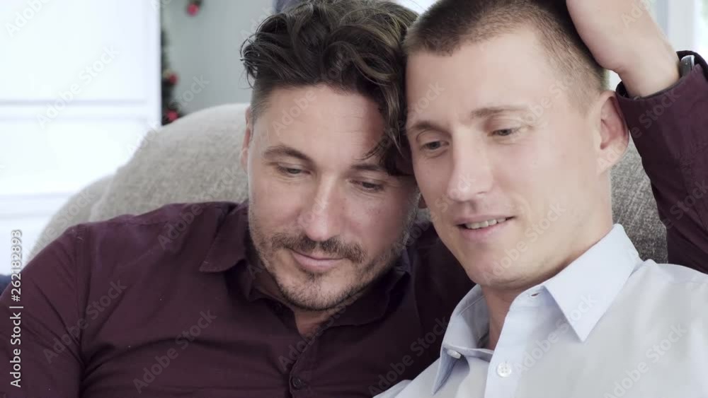 Young gay male couple relax on couch, reading book. Head close together intimate. Happy gay friendship, relationship, pride concept. Young gay male couple relax on couch, reading book. Head close together intimate. Happy gay friendship, relationship, pride concept.