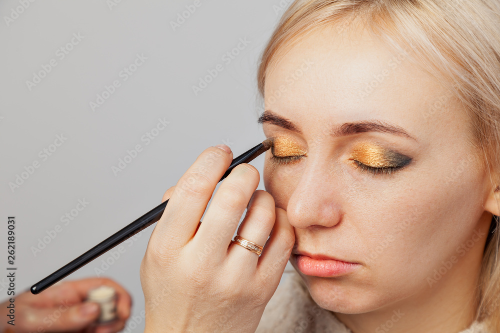Fototapeta premium Visagiste with a brush in one hand putting makeup on the eyes of the model, the other hand pulls the skin of the eyelid with gold and green shadows in east style.