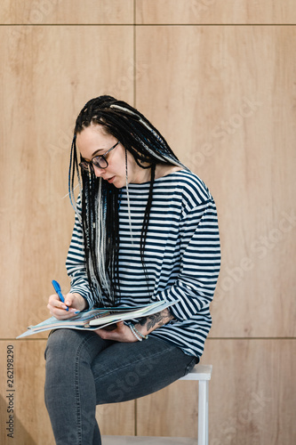 young  smiling dreadlock girl in black glasses making notes in her notebook
