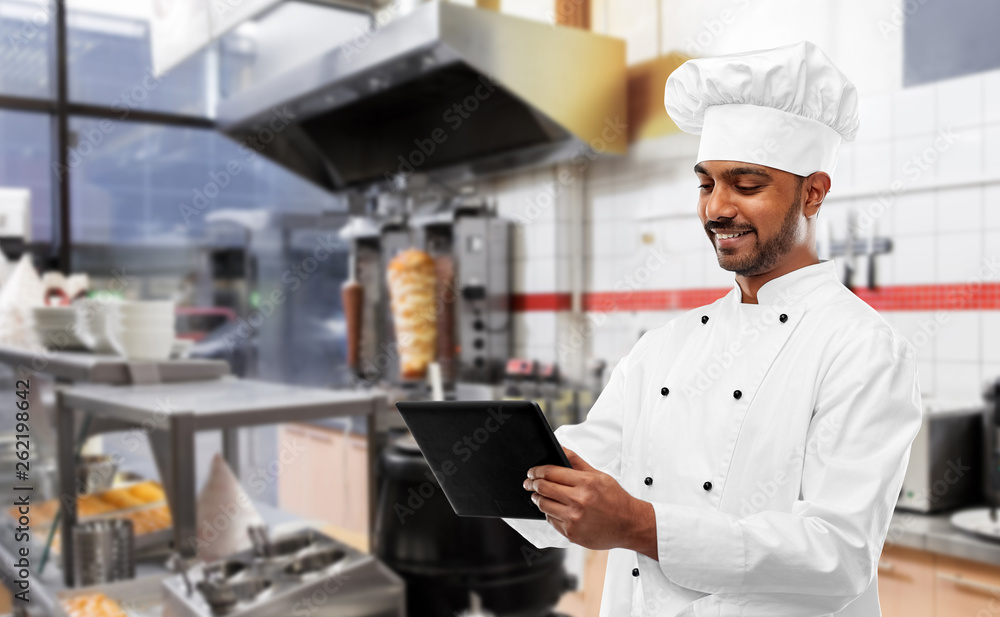 cooking, technology and people concept - happy male indian chef in toque with tablet computer over kebab shop kitchen background