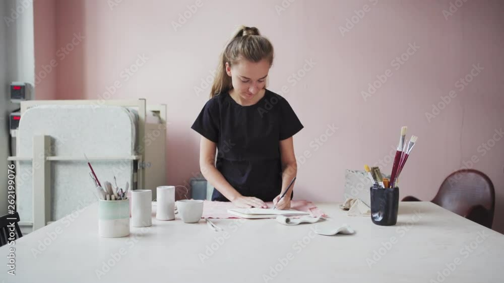 Zoom in medium shot of young female pottery maker sketching at desk in her studio. Unfinished artworks, pieces of clay and mugs with brushesin front of her Zoom in medium shot of young female pottery maker sketching at desk in her studio. Unfinished artworks, pieces of clay and mugs with brushesin front of her