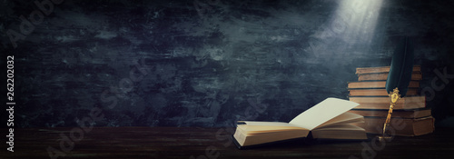 Old feather quill ink pen with inkwell and old books over wooden desk in front of black wall background.  photo