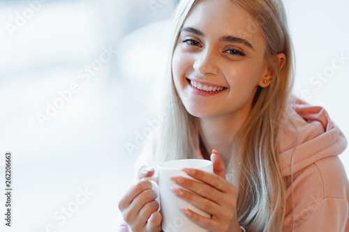 An elf alike, young girl enjoying a cup of coffee or milk while sitting by the window indoors