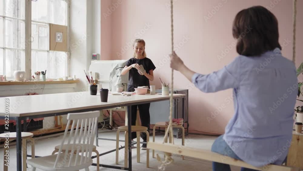 Wide shot of young female ceramic artist making clay artwork at table and talking to her friend relaxing on wooden swing in art studio Wide shot of young female ceramic artist making clay artwork at table and talking to her friend relaxing on wooden swing in art studio