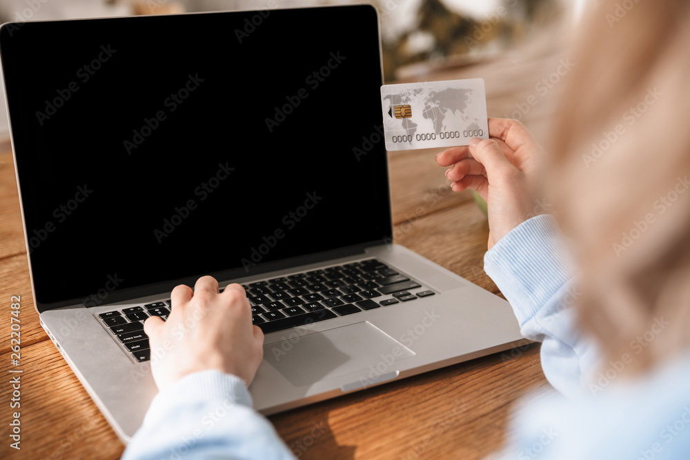 Portrait of attractive blond woman 20s using laptop and credit card while sitting at wooden table indoor - obrazy, fototapety, plakaty 