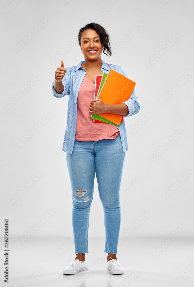 education, school and people concept - happy african american young student woman with notebooks showing thumbs up over grey background education, school and people concept - happy african american young student woman with notebooks showing thumbs up over grey background