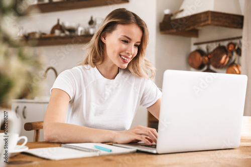Image of european blond woman 20s wearing white t-shirt studying on laptop and writing down notes in apartment