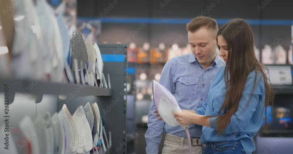 Young married couple chooses an iron to buy in a new home in an appliance store. A woman holds in her hands and looks at the design a man advises more advanced characteristics Young married couple chooses an iron to buy in a new home in an appliance store. A woman holds in her hands and looks at the design a man advises more advanced characteristics