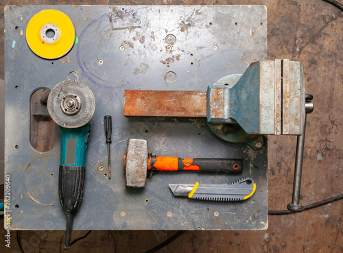 Flat lay view of a workbench with a set of tools consisting of a large heavy vise, angle grinder, screwdriver, cutter, sledgehammer, and a spare cutting disc in a workshop.
