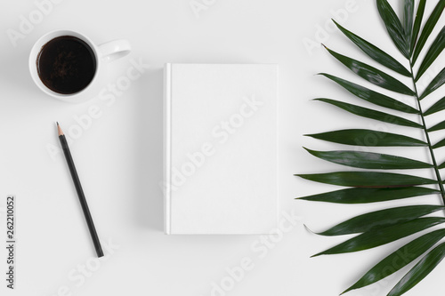 Top view of a white book mockup with workspace accessories and a palm leaf on a white table.