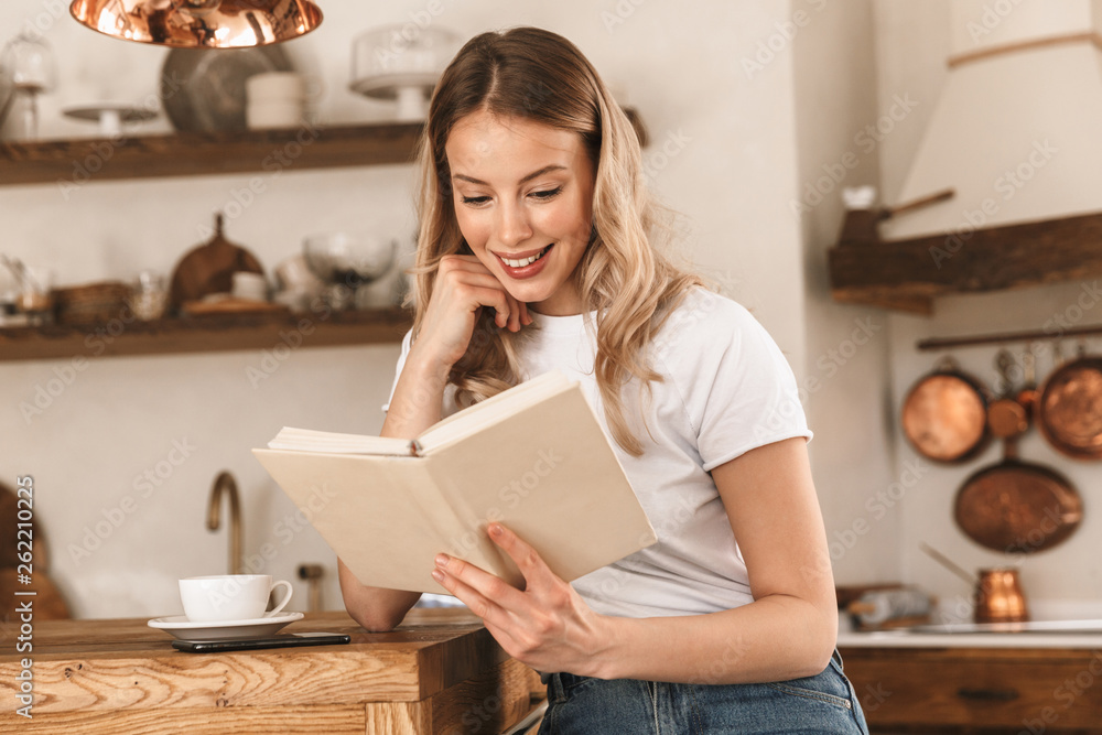 Portrait of young blond woman reading book while standing in stylish wooden kitchen at home Portrait of young blond woman reading book while standing in stylish wooden kitchen at home
