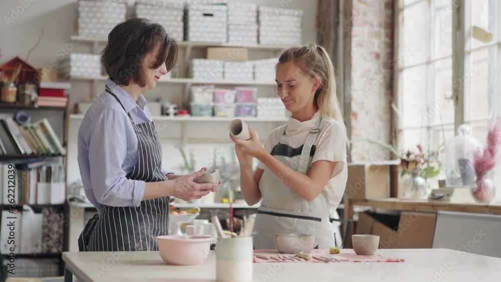 Adult female student bringing young art teacher handmade clay vase and bowl. Teacher showing her how to wipe future artworks with wet sponge Adult female student bringing young art teacher handmade clay vase and bowl. Teacher showing her how to wipe future artworks with wet sponge
