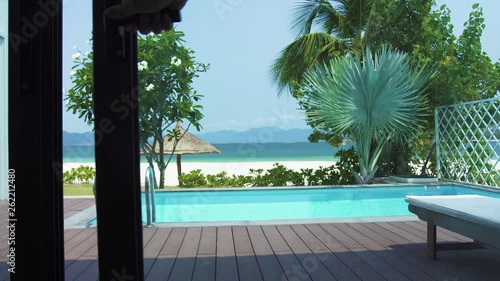 Point of view exit to veranda with swimming pool on sea beach landscape. View from window on swimming pool on luxury villa on sea beach, blue water and palm tree background.