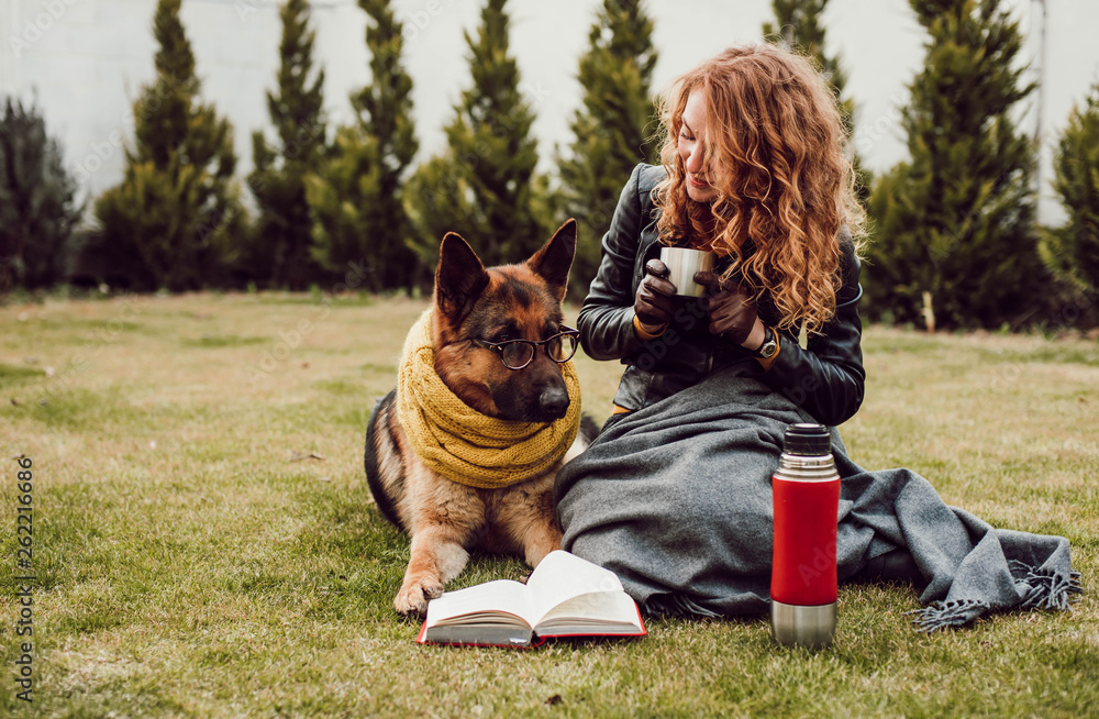 Happy young woman holding thermos cup and sitting with her German shepherd dog on the grass, outdoor Happy young woman holding thermos cup and sitting with her German shepherd dog on the grass, outdoor
